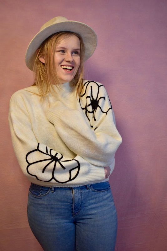 a woman wearing a white sweater with two large embroidered flowers. One is on the right sleeve and the other is on the left shoulder