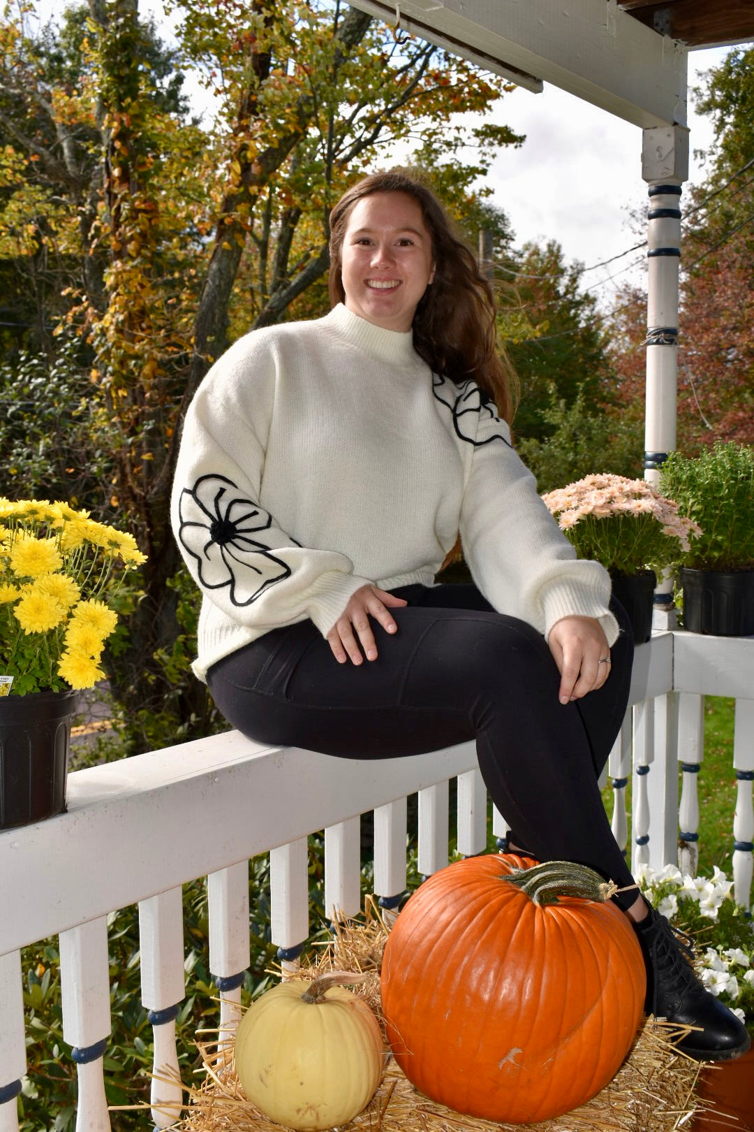 a woman wearing a white sweater with two large embroidered flowers. One is on the right sleeve and the other is on the left shoulder