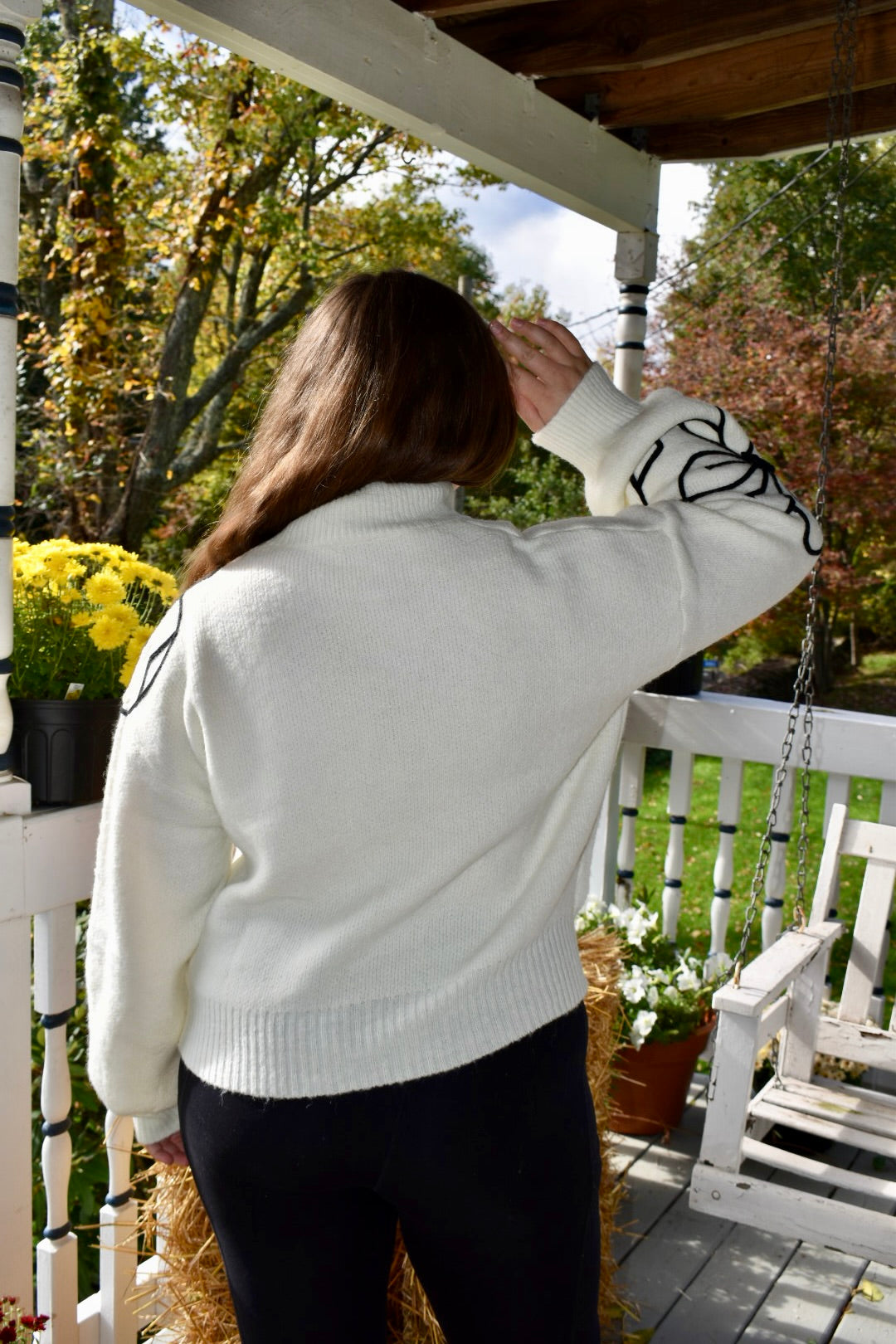 a woman wearing a white sweater with two large embroidered flowers. One is on the right sleeve and the other is on the left shoulder