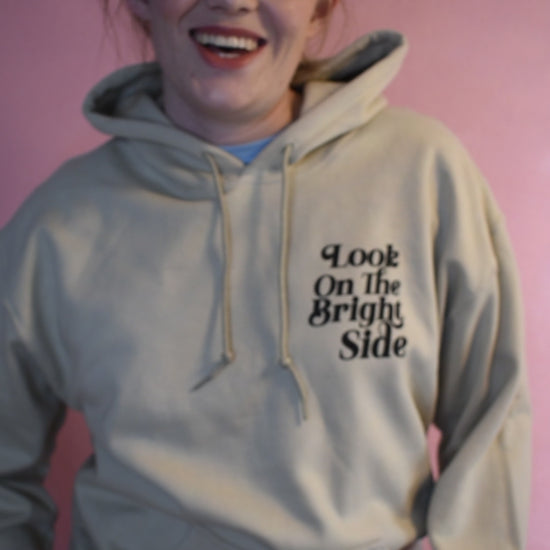 Woman wearing a beige hoodie printed with the words Look On The Bright Side. She is talking about why she likes it